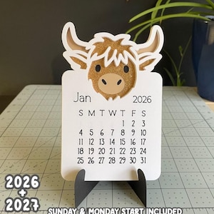 May include: A white desk calendar for January 2026, featuring a Highland cow design with glitter accents. The calendar includes the days of the month and is supported by a black stand. Text at the bottom reads "SUNDAY & MONDAY START INCLUDED."