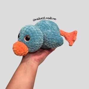 May include: A crocheted blue duck plush toy with orange beak and feet. The duck has large black eyes and is held in a hand. The text "@midwest.made.mn" is visible in the image.