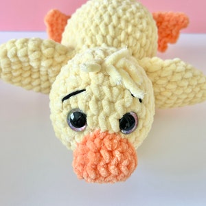 May include: A crocheted yellow duck plush toy with orange beak and feet. The duck has large black eyes with purple pupils.