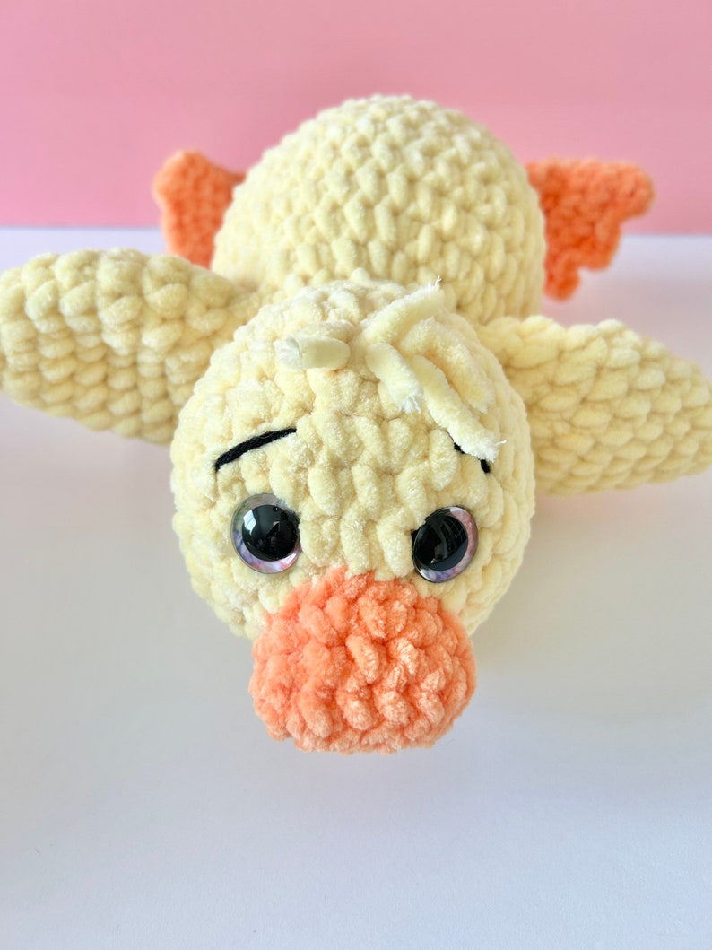 May include: A crocheted yellow duck plush toy with orange beak and feet. The duck has large black eyes with purple pupils.