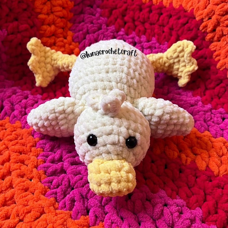 May include: A white crocheted duck with yellow feet and beak, lying on a colorful crocheted blanket. The duck has black eyes and a fluffy texture. The blanket is made of a variety of colors, including pink, orange, and red.