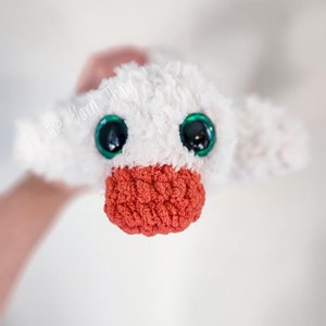 May include: A white crocheted plush toy with green eyes and a red crocheted beak. The toy is being held by a hand.