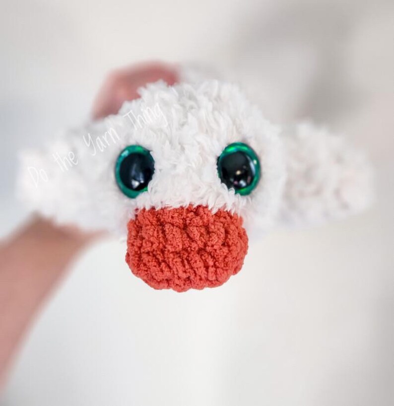 May include: A white crocheted plush toy with green eyes and a red crocheted beak. The toy is being held by a hand.