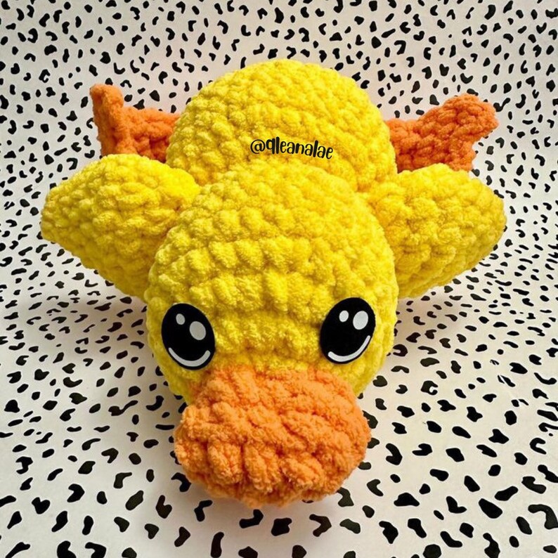 May include: A crocheted yellow duck plush toy with orange accents and black eyes. The toy is sitting on a black and white patterned surface. The text "@gleanaiae" is visible on the toy.