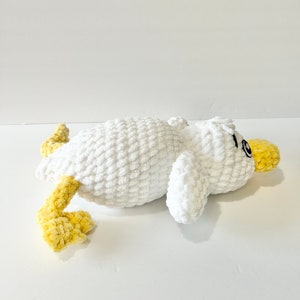 May include: A white crocheted plush toy duck with yellow crocheted legs and a black eye. The duck is lying on its side with its head turned to the side.