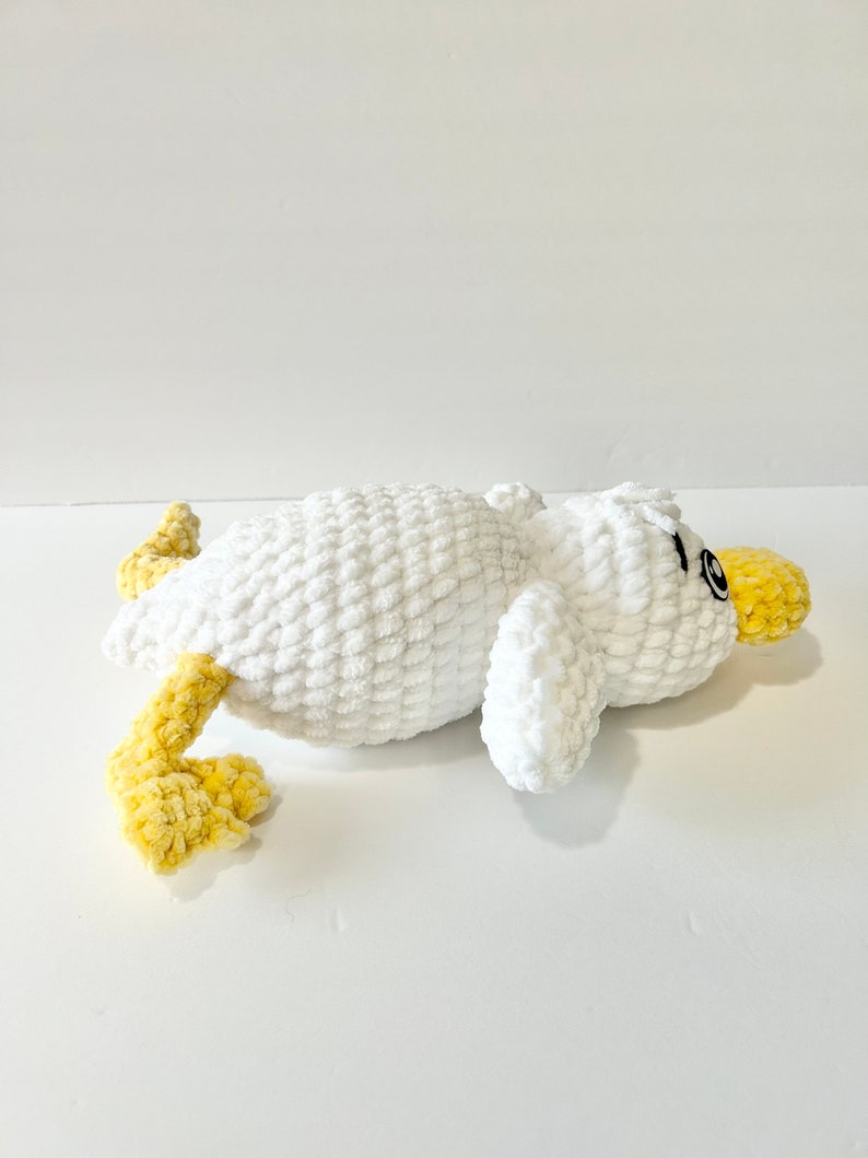 May include: A white crocheted plush toy duck with yellow crocheted legs and a black eye. The duck is lying on its side with its head turned to the side.