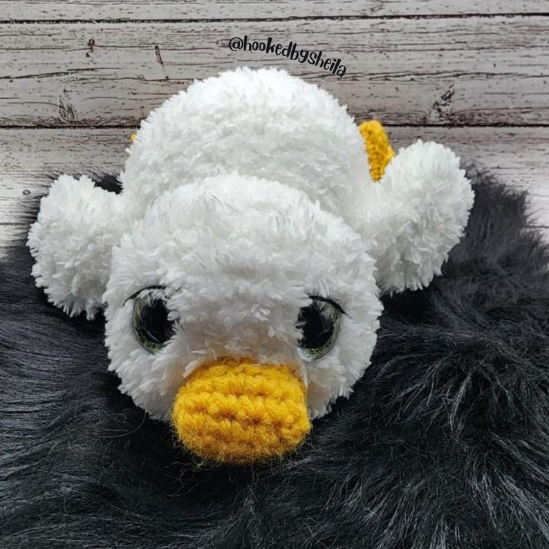 May include: A white crocheted duck with yellow beak and feet, lying on a black fuzzy surface. The duck has large black eyes with green pupils. The text "@hookedbysheila" is visible in the background.