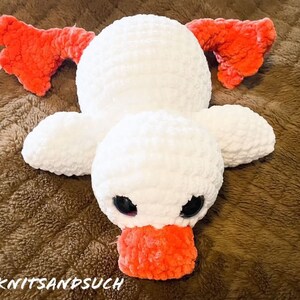 May include: A white crocheted duck plush toy with orange felt wings and beak. The duck is lying on a brown fuzzy surface.