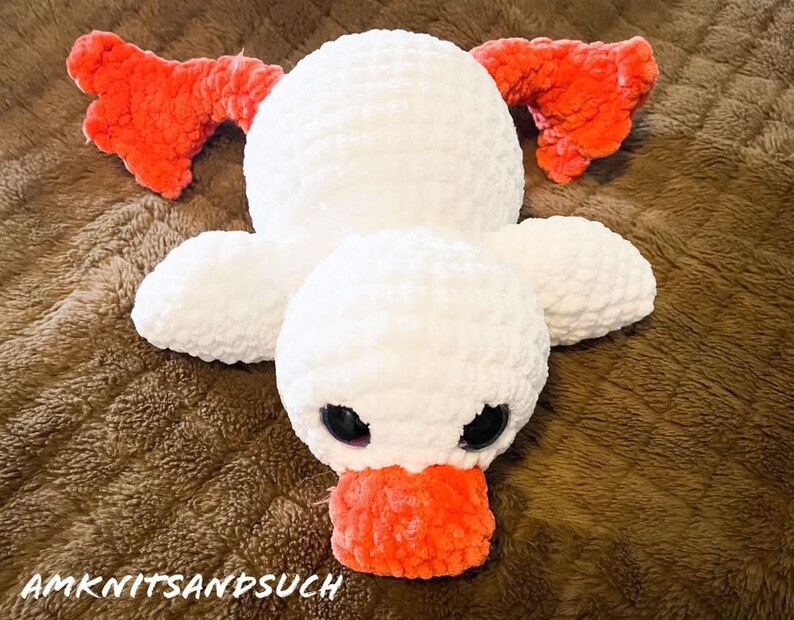 May include: A white crocheted duck plush toy with orange felt wings and beak. The duck is lying on a brown fuzzy surface.