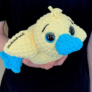 May include: A crocheted yellow duck plush toy with blue eyes, a blue beak, and a blue tail fin. The toy is being held by a person's hand.