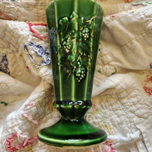 May include: A green ceramic vase with a raised design of grape vines and leaves. The vase has a flared top and a wide base.