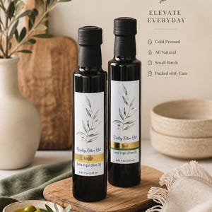 May include: Two dark glass bottles of Valley Olive Oil, one labeled "Lemon" and the other "Basil." Each bottle has a black cap and a white label with a botanical design. The bottles sit on a wooden board with a small dish of olives and a sprig of leaves.