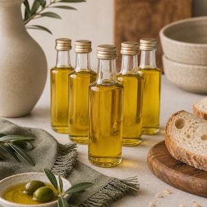 May include: Five small glass bottles filled with golden olive oil, each topped with a gold cap, are arranged on a light surface. A small dish with olives and olive oil, along with a sprig of olive leaves, is in the foreground. Sliced bread and a vase are also present.