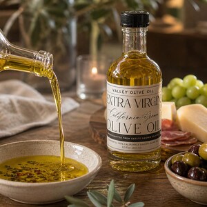 May include: A bottle of Valley Olive Oil Extra Virgin California Green Olive Oil, 8.5 fl oz (250 ml), with a black cap. Olive oil is being poured into a small bowl. Olives, cheese, and grapes are also present.