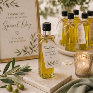 May include: A small glass bottle filled with yellow liquid, tied with twine and a tag that reads "Thank You". The bottle sits on a square stone, with a sign that says "Thank You for Sharing Our Special Day". A lit candle and olive branches are also present.
