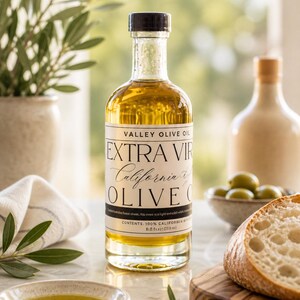 May include: A clear glass bottle of Valley Olive Oil with a black cap, labeled "Extra Virgin California Olive Oil." The bottle is filled with golden olive oil. A small bowl of olive oil, olives, and bread are also in the image.