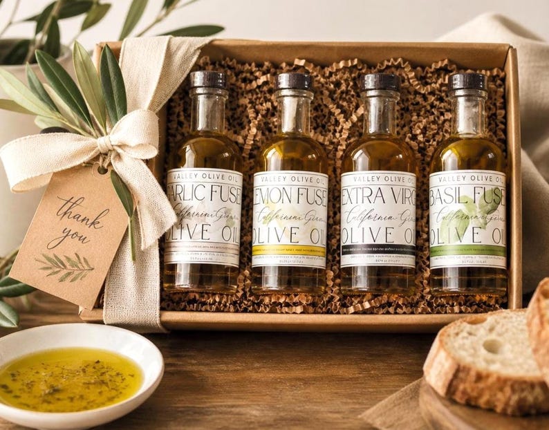 May include: A gift box containing four olive oil bottles with different flavors: Garlic Fuse, Lemon Fuse, Extra Virgin, and Basil Fuse. Each bottle has a label with the flavor name. A small bowl of olive oil and bread slices are also present.