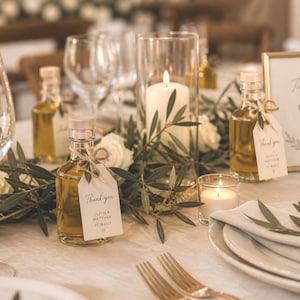 May include: A wedding reception table setting with clear wine glasses, plates, and gold-colored forks. Small glass bottles of olive oil with tags that say "Thank you" are placed on the table, along with candles and floral arrangements.