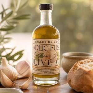 May include: A clear glass bottle of Valley Olive Oil Garlic Fused California Garlic Olive Oil, 8.5 fl oz (250 ml), with a black cap. The bottle is on a wooden surface with garlic cloves, a small bowl of oil, and a loaf of bread.