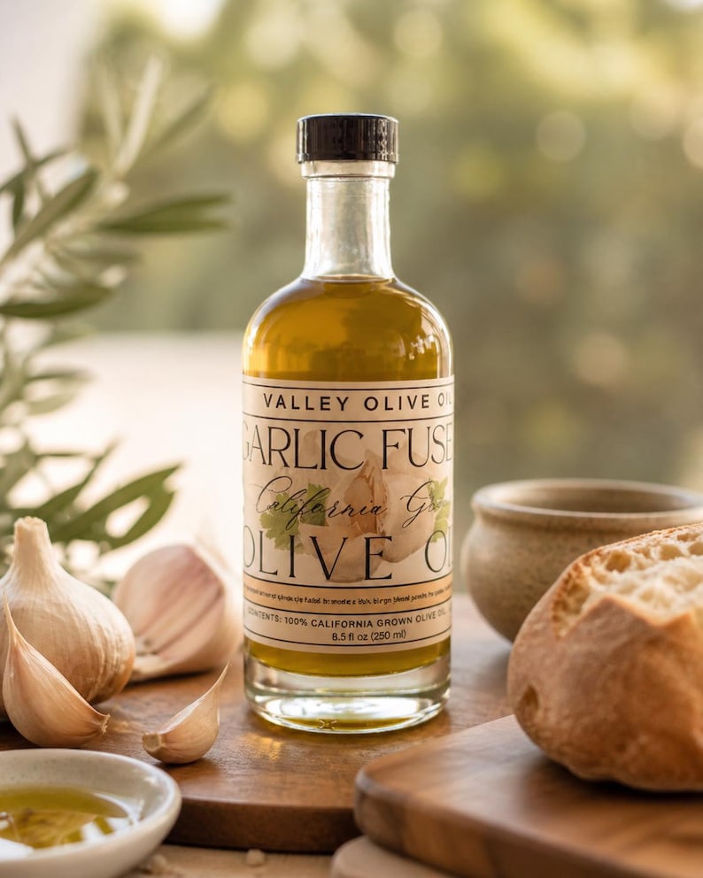 May include: A clear glass bottle of Valley Olive Oil Garlic Fused California Garlic Olive Oil, 8.5 fl oz (250 ml), with a black cap. The bottle is on a wooden surface with garlic cloves, a small bowl of oil, and a loaf of bread.