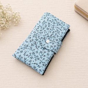 May include: A rectangular fabric wallet in light blue featuring a black branch and twig pattern. The wallet has a fold-over flap secured with a single white snap button. A glimpse of a black and white polka dot fabric is visible on the side edge.
