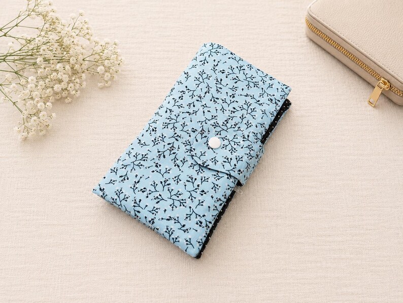 May include: A rectangular fabric wallet in light blue featuring a black branch and twig pattern. The wallet has a fold-over flap secured with a single white snap button. A glimpse of a black and white polka dot fabric is visible on the side edge.