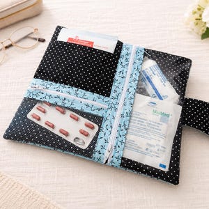 May include: A black fabric pouch with white polka dots featuring a light blue floral trim and two clear vinyl pockets. The pouch contains a blister pack of red capsules and medical supplies, secured with a snap-button flap closure.