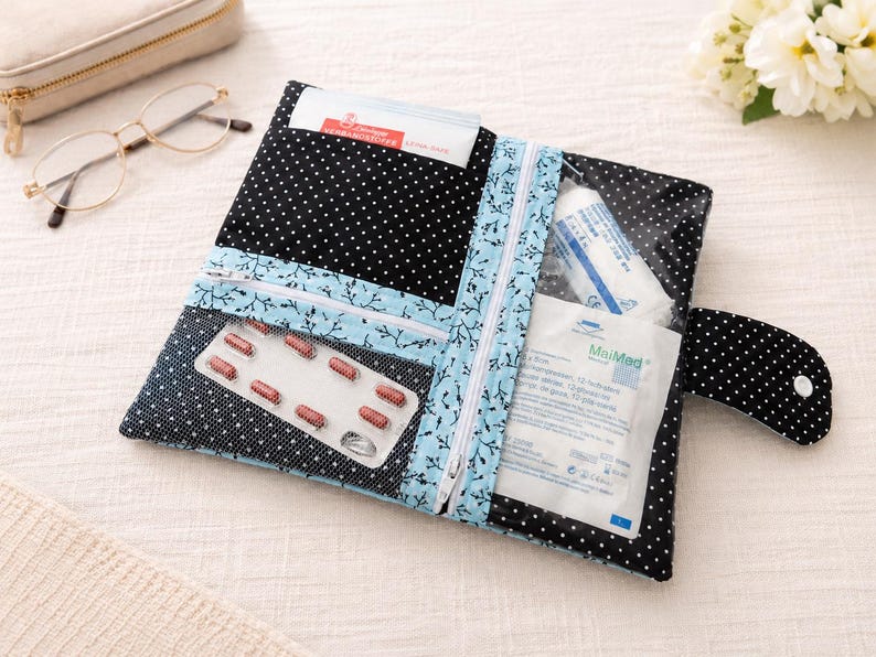 May include: A black fabric pouch with white polka dots featuring a light blue floral trim and two clear vinyl pockets. The pouch contains a blister pack of red capsules and medical supplies, secured with a snap-button flap closure.