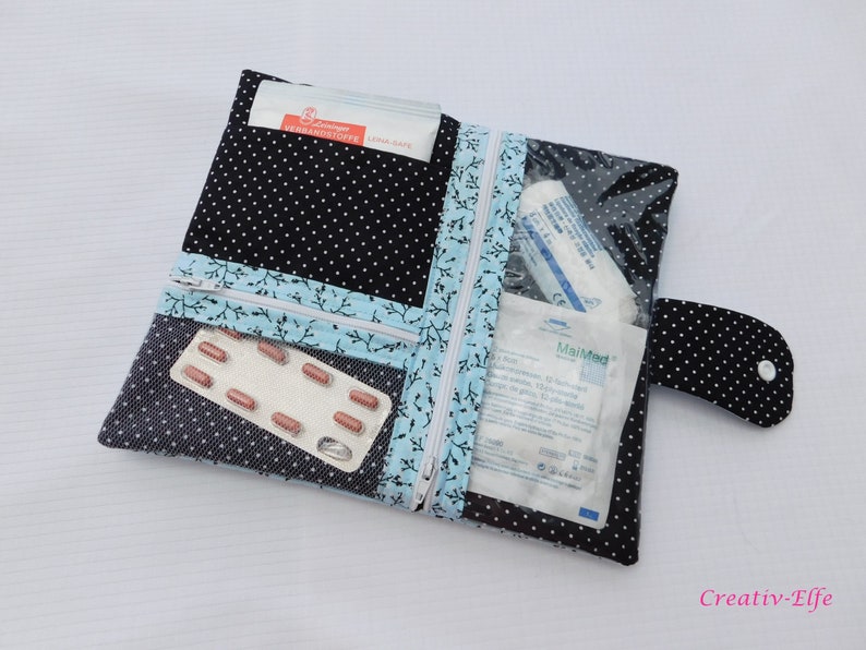 May include: A black fabric pouch with white polka dots featuring a light blue trim with a black branch pattern. The pouch has two zippered compartments, one with a clear plastic window, containing medical supplies like bandages and a blister pack.