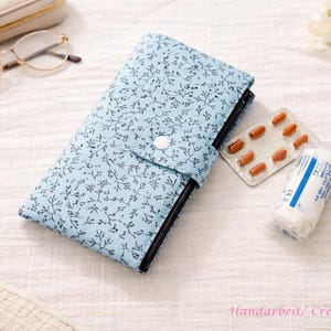 May include: A light blue fabric wallet featuring a dark blue branch and twig pattern with a single white snap closure. The wallet is positioned next to a blister pack of ten brown capsules and a small roll of white medical tape with blue text.