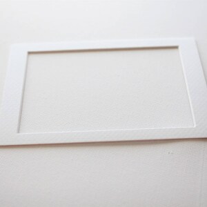 Puede incluir: Una cartulina blanca con una abertura rectangular en el centro. La cartulina es texturizada y tiene un borde ligeramente elevado.