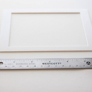 Puede incluir: Una cartulina blanca con una abertura rectangular en el centro. Una regla se coloca debajo de la cartulina para mostrar el tama&ntilde;o. La regla est&aacute; marcada en pulgadas.
