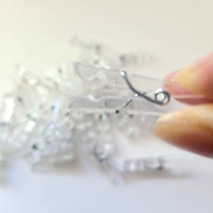 May include: A close-up of a pile of clear plastic curtain clips with a silver metal hook. The clips are used to hang curtains or drapes.