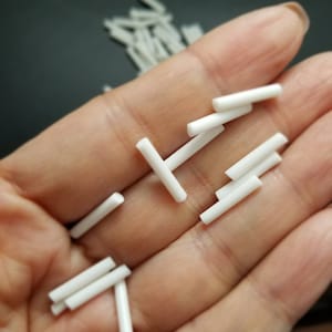 May include: A close-up of a hand holding a pile of small white ceramic rods. The rods are about 1 inch long and have a smooth, rounded surface.