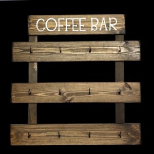 May include: A wall-mounted wooden coffee bar organizer featuring three horizontal planks with a dark brown wood grain finish. Each plank is equipped with four black metal hooks for hanging items. The top header board displays the white text COFFEE BAR.