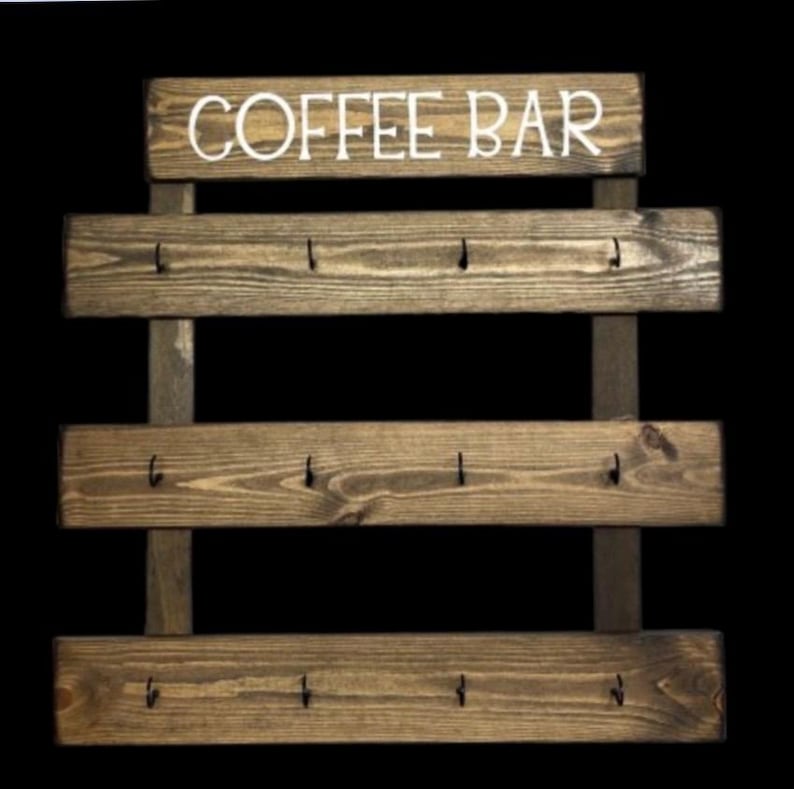 May include: A wall-mounted wooden coffee bar organizer featuring three horizontal planks with a dark brown wood grain finish. Each plank is equipped with four black metal hooks for hanging items. The top header board displays the white text COFFEE BAR.