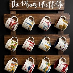 May include: A dark wood wall-mounted mug rack with three horizontal slats and black metal hooks. The top board features the white text "the Places We'll Go". Twelve ceramic mugs are hanging, each decorated with a colorful illustration of a global city including Kyoto, New York City, Paris, Rome, Mexico City, London, Dubai, Seoul, Barcelona, Tokyo, Cape Town, and Toronto.