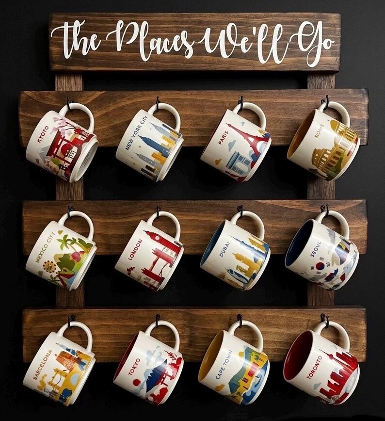 May include: A dark wood wall-mounted mug rack with three horizontal slats and black metal hooks. The top board features the white text "the Places We'll Go". Twelve ceramic mugs are hanging, each decorated with a colorful illustration of a global city including Kyoto, New York City, Paris, Rome, Mexico City, London, Dubai, Seoul, Barcelona, Tokyo, Cape Town, and Toronto.
