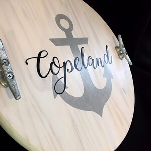 May include: A round light-colored wooden serving tray featuring a large gray anchor graphic in the center. The word Copeland is printed in black cursive script over the anchor. Two silver-toned metal boat cleats are attached as handles.
