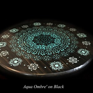 May include: A round wooden table top with a teal and black mandala design. The design features a large central mandala surrounded by smaller mandalas. The table top is painted black with a teal ombre effect.