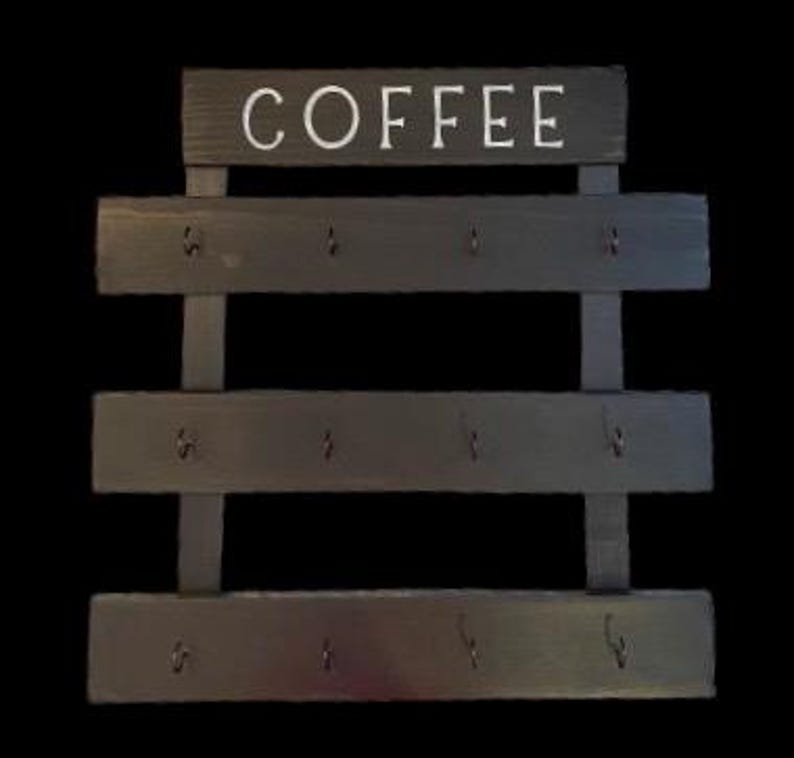 May include: A dark gray wooden wall-mounted rack featuring the word COFFEE in white capital letters at the top. The rack consists of three horizontal wooden slats, each equipped with four small metal hooks for hanging items, totaling twelve hooks.