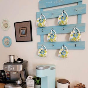 May include: A light blue wooden wall-mounted rack with the word coffee in white script, featuring black metal hooks holding six white ceramic mugs decorated with blue and green floral patterns, displayed above a coffee station with two machines.