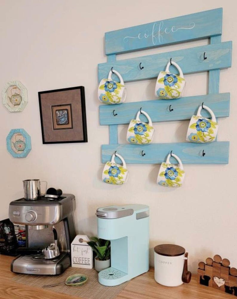 May include: A light blue wooden wall-mounted rack with the word coffee in white script, featuring black metal hooks holding six white ceramic mugs decorated with blue and green floral patterns, displayed above a coffee station with two machines.