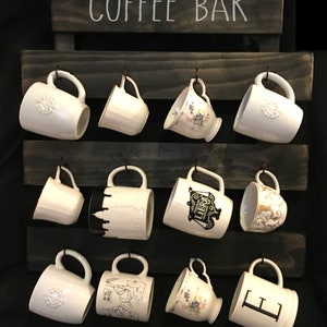 May include: A dark-stained wooden wall-mounted rack with the text COFFEE BAR printed at the top. The rack features three horizontal slats with twelve metal hooks, each holding a white ceramic mug with various designs, patterns, and letters.