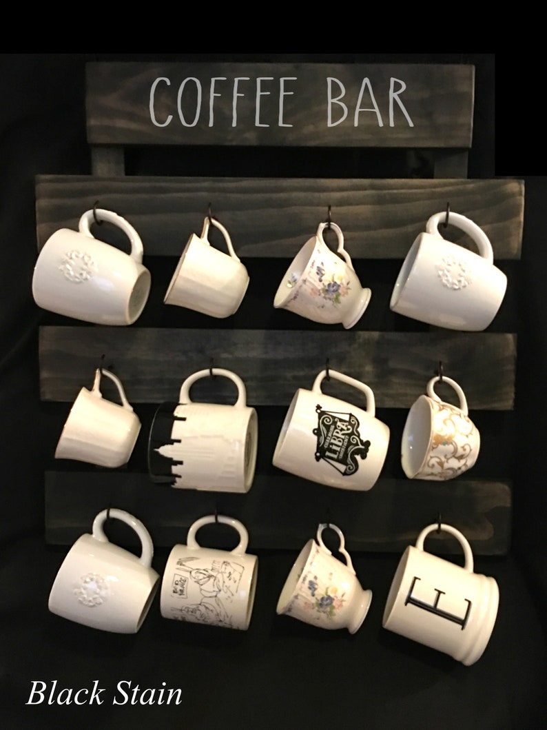 May include: A dark-stained wooden wall-mounted rack with the text COFFEE BAR printed at the top. The rack features three horizontal slats with twelve metal hooks, each holding a white ceramic mug with various designs, patterns, and letters.