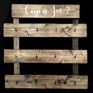 May include: A dark stained wooden wall rack featuring three horizontal planks attached to two vertical supports. Each plank has four black metal hooks. The top plank displays the white cursive text "Cup a Joe ?" printed across the center.