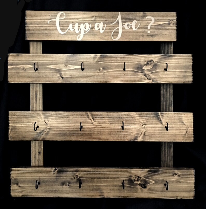 May include: A dark stained wooden wall rack featuring three horizontal planks attached to two vertical supports. Each plank has four black metal hooks. The top plank displays the white cursive text "Cup a Joe ?" printed across the center.