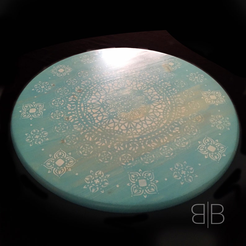 May include: A round, turquoise table top with a white mandala design. The design features intricate details and a faded center.