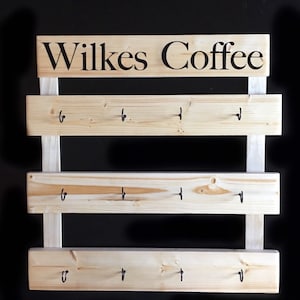 May include: A light-colored wooden wall rack featuring three horizontal planks, each equipped with four black metal hooks. The top plank displays the black text Wilkes Coffee in a serif font. The structure is held together by two vertical wooden supports.