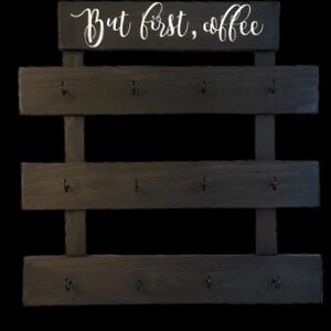 May include: A dark wood wall-mounted rack featuring three horizontal slats, each equipped with four black metal hooks. The top section displays the white cursive text "But first, coffee" printed on a rectangular wooden board.